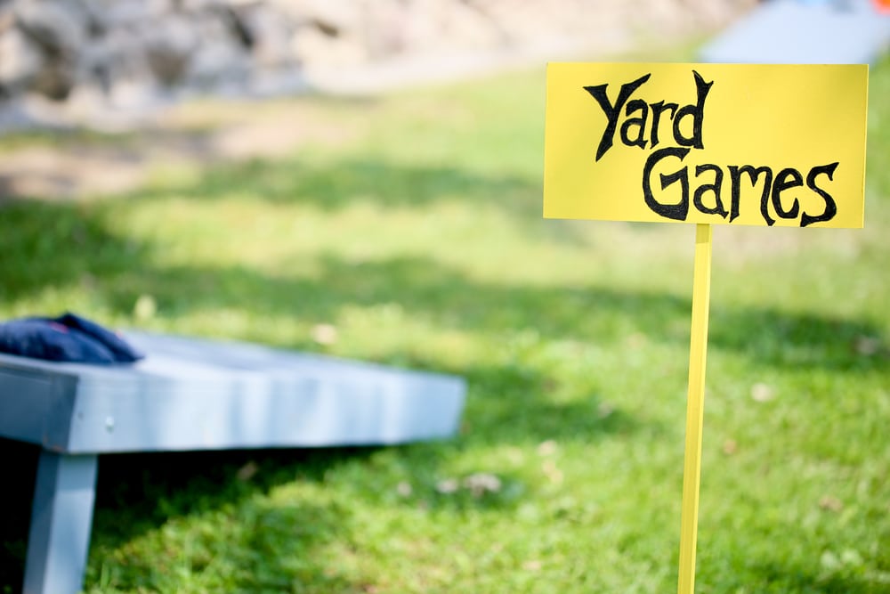 Yellow Yard Games Sign on Stick With Cornhole Board sitting on grass in background